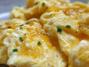 Close-up of perfectly fluffy scrambled eggs, glistening with butter and topped with fresh chives and black pepper.