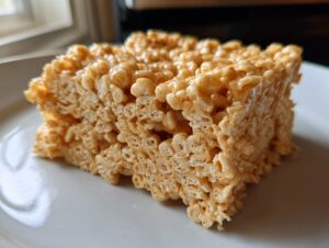 A thick, gooey square of peanut butter rice krispie treats sitting on a white plate.