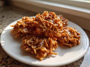 A close-up of several shiny, caramel-colored haystack cookies resting on a white plate near a window.