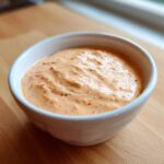 Close-up of creamy, orange-hued homemade Big Mac Sauce speckled with red relish bits in a white bowl.