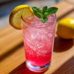 A tall glass of honey lemon pink soda filled with ice, garnished with a lemon slice and fresh mint leaves.