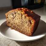 A thick, moist slice of sugar free banana bread with visible nuts, sitting on a white plate.