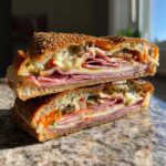 A stacked, halved Muffuletta sandwich showcasing layers of cured meats, cheese, and olive salad on a sesame seed bun.