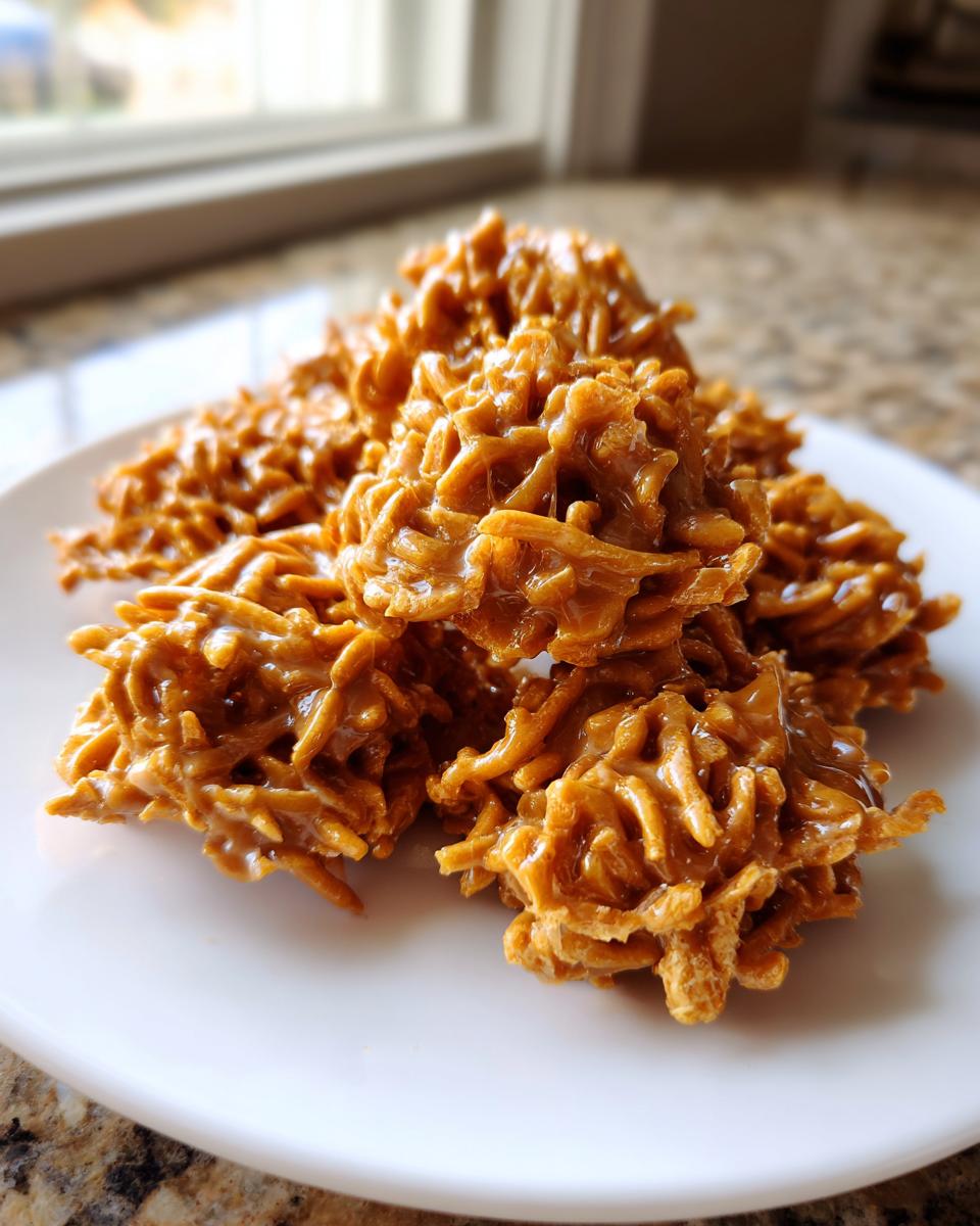 A pile of glossy, caramel-colored haystack cookies made with chow mein noodles on a white plate.
