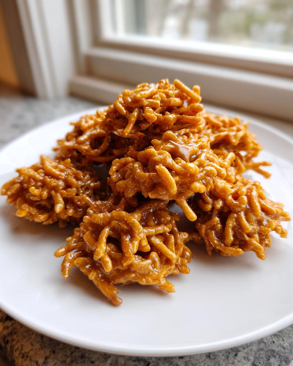 A pile of glossy, peanut butter-colored haystack cookies resting on a white plate near a window.