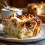 A golden slice of old-fashioned bread pudding being drizzled with a creamy sauce from a spoon.
