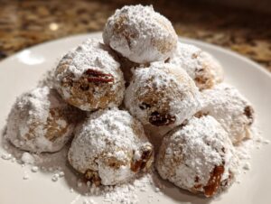 A pile of freshly baked Pecan Snowball Cookies generously coated in powdered sugar, with visible pecan pieces.