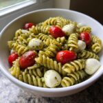 A white bowl filled with pesto pasta salad featuring rotini pasta, cherry tomatoes, and small mozzarella balls.