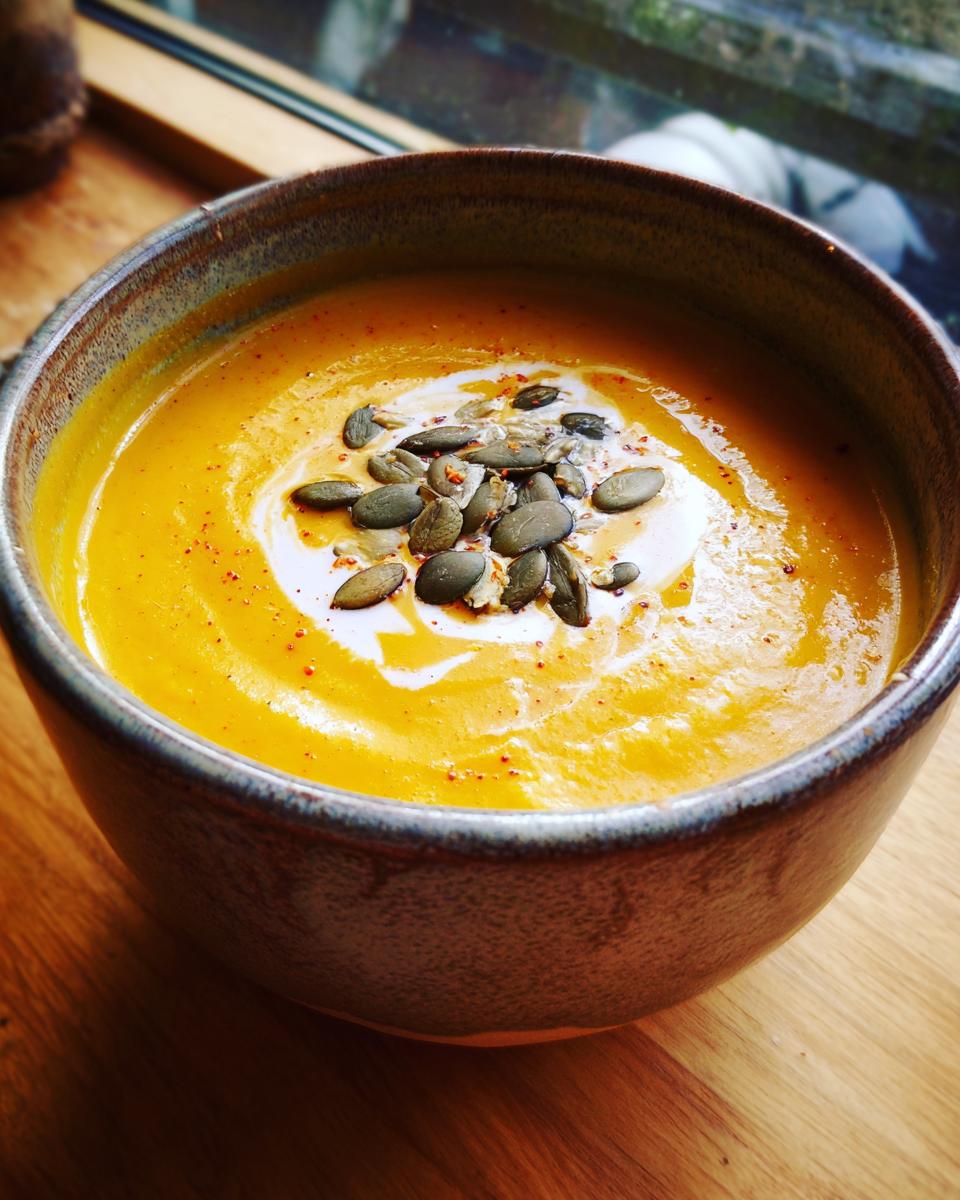 A close-up of a bowl of vibrant roasted pumpkin soup, topped with cream, pumpkin seeds, and a sprinkle of paprika.