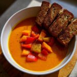 A vibrant bowl of roasted vegetable soup topped with colorful bell pepper chunks and served with toasted bread slices.