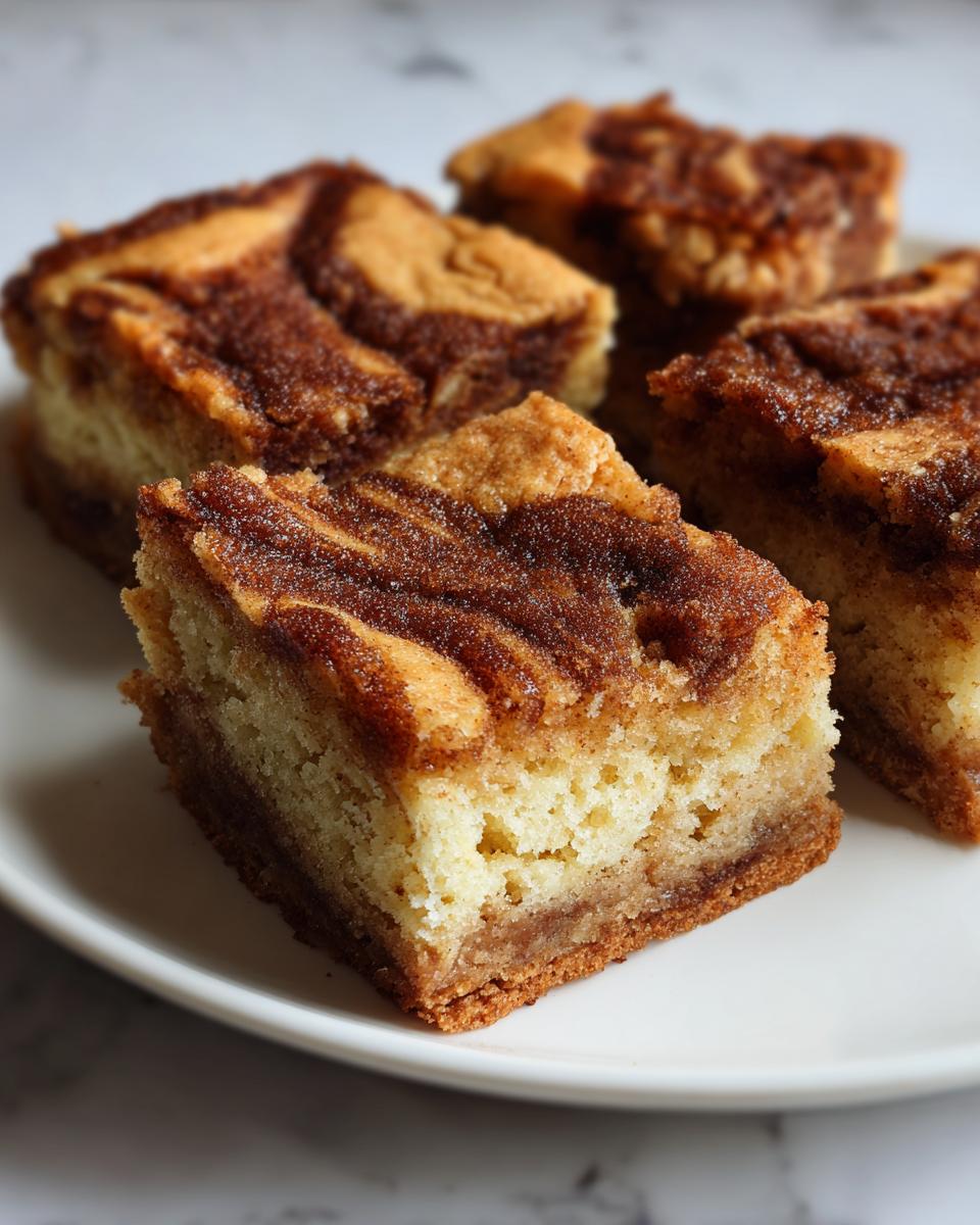 Four square-cut snickerdoodle blondies featuring a rich cinnamon swirl topping on a white plate.
