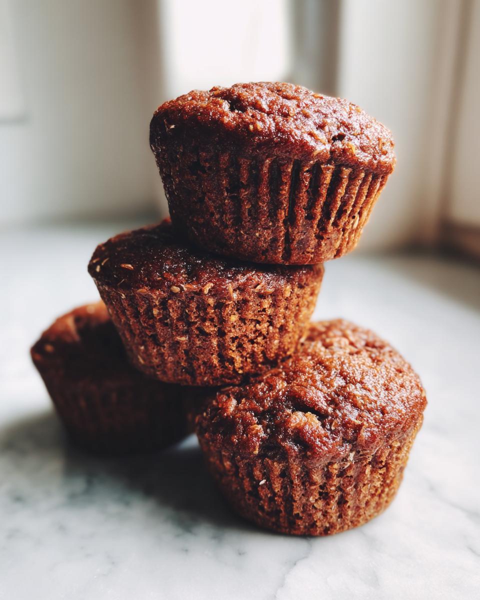 A small stack of four dark, moist bran muffins sitting on a white marble surface.