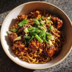 A close-up overhead shot of sticky garlic chicken noodles topped with sesame seeds and green onions.