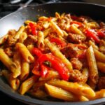 Close-up of a pan filled with sweet pepper and sausage pasta, featuring penne pasta, sliced red peppers, and crumbled sausage in a rich sauce.