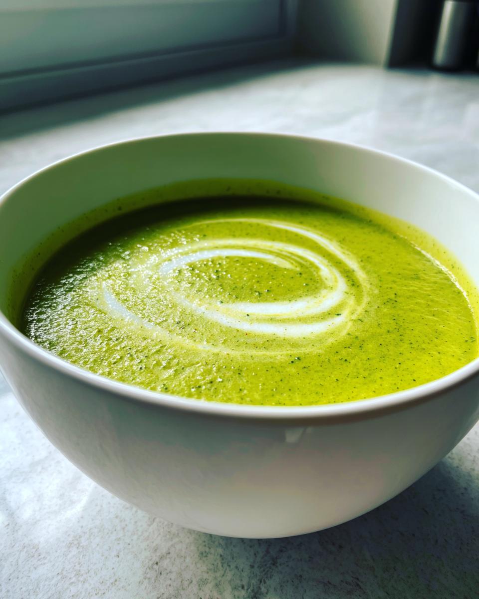 A close-up of bright green zucchini soup topped with a white cream swirl in a white bowl.