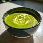 Close-up of a bowl of bright green zucchini soup topped with a decorative swirl of white cream.