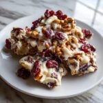 A close-up of white chocolate cranberry pecan clusters piled on a white plate, showcasing the sweet and nutty ingredients.