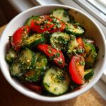 Close-up of a fresh cucumber tomato salad tossed in a bright herb dressing, served in a white bowl.