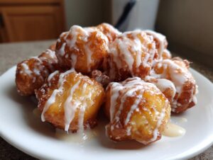 A close-up of golden brown apple fritter bites piled on a white plate, drizzled generously with white vanilla glaze.