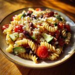 A vibrant bowl of greek pasta salad featuring fusilli, cherry tomatoes, cucumber, olives, and crumbled feta cheese.