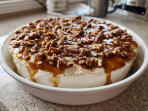 A rich pecan pie dip topped with glossy caramel sauce and chopped pecans, served in a white bowl.