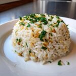 A mound of white rice pilaf, studded with toasted nuts and topped with fresh chopped parsley, served on a white plate.