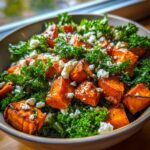 A close-up of a vibrant sweet potato salad featuring roasted orange sweet potato cubes mixed with dark green kale and topped with crumbled white cheese.