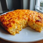 A golden-brown, baked cream cheese corn casserole served on a white plate with a piece already scooped out.