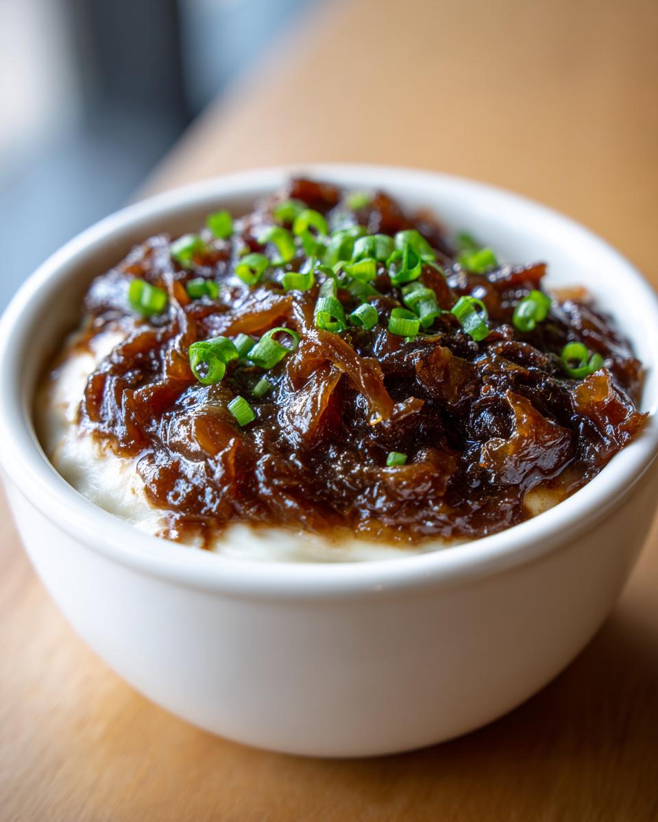 Close-up of a white bowl filled with creamy caramelized onion dip, topped with dark onions and fresh green chives.