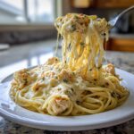 A fork lifts a large portion of creamy Monterey chicken spaghetti, showing long, stretchy cheese pulls over the main serving.