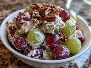 A white bowl filled with creamy chicken salad chick grape salad, featuring red and green grapes and topped with pecans.