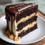A tall slice of rich chocolate peanut butter cake with three layers, peanut butter frosting, and chocolate ganache drip.