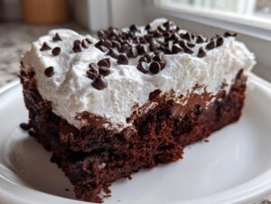 A close-up of a moist slice of chocolate poke cake topped with thick white frosting and mini chocolate chips.