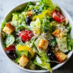 A vibrant close-up of a bowl filled with copycat Olive Garden salad, featuring romaine lettuce, croutons, olives, tomatoes, and grated cheese.
