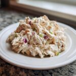 A mound of creamy cottage cheese chicken salad featuring shredded chicken, red onion, and celery on a white plate.