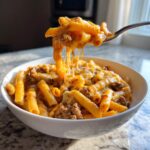 A fork lifts a cheesy, saucy portion of creamy beef pasta out of a white bowl, showing long cheese pulls.