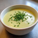 A close-up of creamy, pale green celery soup topped generously with chopped fresh chives in a white bowl.