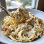 A fork lifting a bite of creamy chicken alfredo pasta, rich with white sauce, chicken pieces, and grated cheese.