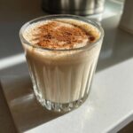 A close-up of a glass filled with creamy coquito, topped generously with ground cinnamon.
