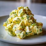 A mound of creamy, chunky egg salad recipe mixed with celery and chives, served on a white plate.