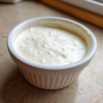 Close-up of thick, creamy garlic sauce in a small white fluted ramekin on a countertop.