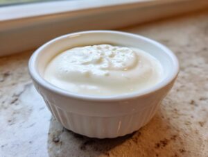 Close-up of a small white ramekin filled with thick, creamy garlic sauce, resting on a speckled countertop.