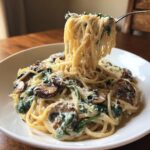 A fork lifts a generous swirl of creamy mushroom pasta, showing spaghetti coated in sauce with sautéed mushrooms and spinach.