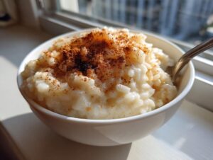 Close-up of a bowl of creamy rice pudding topped generously with ground cinnamon, sitting by a sunlit window.