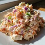 A close-up, bright photo of creamy seafood salad mixed with shredded carrots, red onion, and green onion pieces.