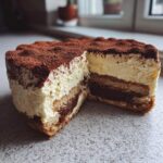 A close-up of a slice removed from a tiramisu cheesecake showing layers of cream, coffee-soaked cake, and cocoa dusting.