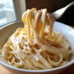 A fork lifting fettuccine coated in rich vegan alfredo sauce from a white bowl.