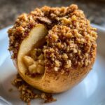 A close-up of a perfectly baked apple with a thick, golden-brown oat crumble topping, served on a white plate.