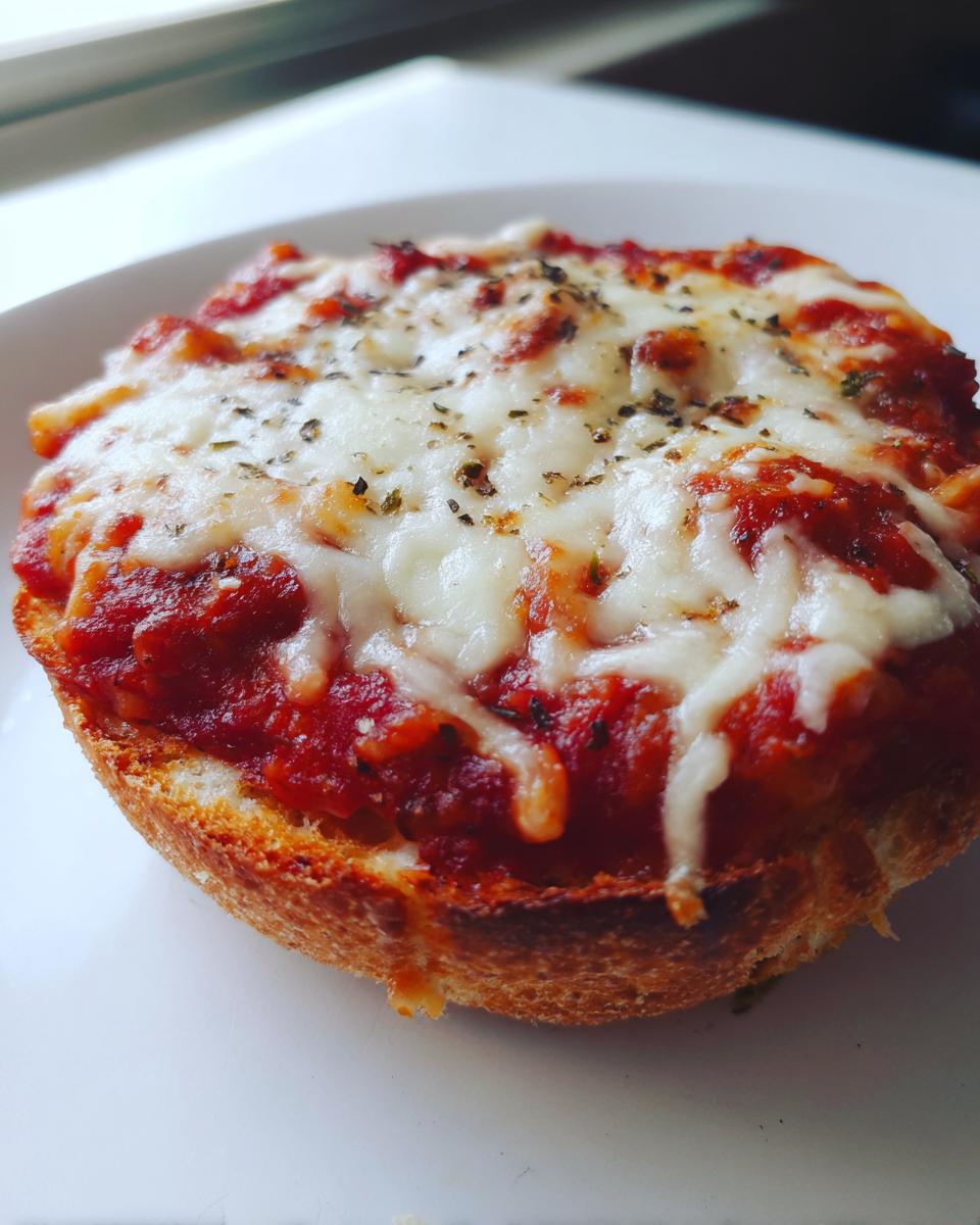 Close-up of a toasted english muffin pizza half topped with rich tomato sauce, melted mozzarella, and dried herbs.
