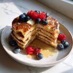 A tall stack of fluffy almond flour pancakes, cut to show the texture, topped with butter, syrup, blueberries, and raspberries.